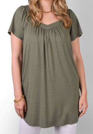 Vrouw draagt een olijfgroene tuniek met korte mouwen en een geraffineerde V-hals, een gouden armband en ketting, gecombineerd met een witte broek.