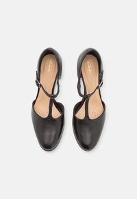 Escarpins en cuir noir avec un design à bride en T, bout arrondi et petit talon. Intérieur avec une doublure lisse et des détails perforés.