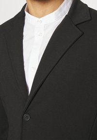 Sort blazer med en glat finish, der har en spids slag, enkelt knaplukning og en hvid skjorte under.