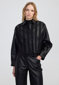 Czarna skórzana kurtka typu cropped z pionowymi paskami o matowej i błyszczącej fakturze, z kołnierzykiem i zapięciem na guziki.