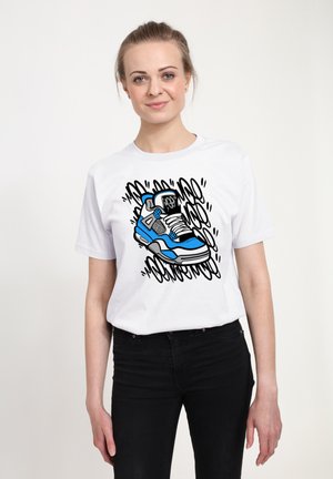 Młoda kobieta ubrana w białą koszulkę z niebiesko-czarną grafiką trampka i czarne spodnie, stojąca na tle gładkiego jasnego tła.