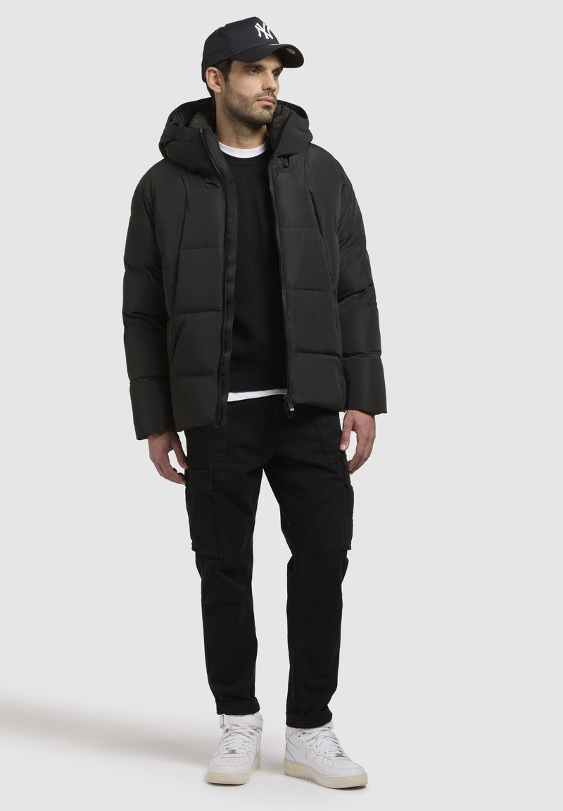 Mann in schwarzer Daunenjacke, schwarzen Cargo-Hosen, weißen Sneakers und einer marineblauen Kappe mit weißem Logo, der vor einem schlichten Hintergrund steht.