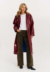 Burgundy umelá koža dlhý kabát s viditeľným šitím, nosený cez biely tričko a hnedé zvonové nohavice s leopardím vzorom, doplnený čiernymi čižmami.