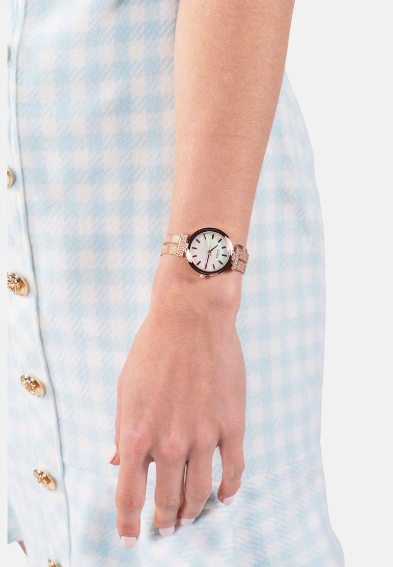 Rožnato zlat ure s okroglo številčnico, svetlo zelenim dialom in strukturo mrežastega pasu. Oblečeno na roki proti svetlo modri karirasti podlagi.