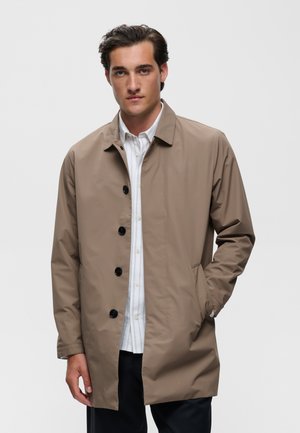 Jeune homme portant un manteau beige boutonné sur une chemise blanche à rayures, une main dans la poche, debout devant un fond blanc uni.