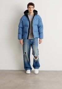 Light blue puffer jacket with black accents, zippered front, and hood, worn over a gray shirt and distressed blue jeans. White sneakers.