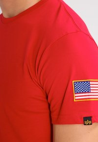 Camisa roja de mangas cortas usada por una persona con un parche de la bandera estadounidense en el brazo superior y una pequeña etiqueta negra y amarilla en el dobladillo de la manga.