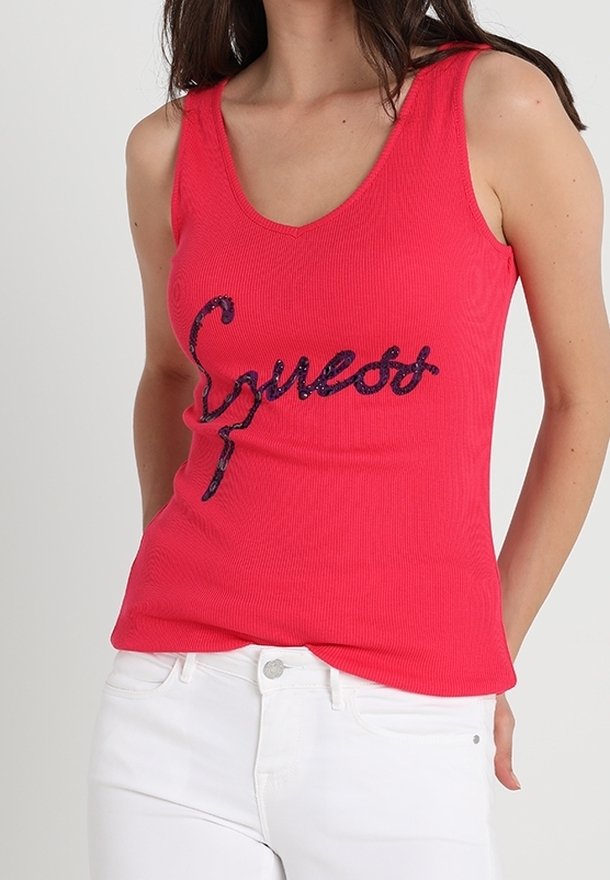 Femme portant un haut sans manches rouge avec "Guess" écrit en sequins foncés, associé à un pantalon blanc sur fond uni.