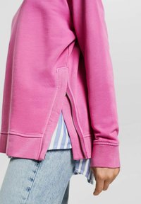 Persona che indossa una maglietta a maniche lunghe rosa con zip laterale sopra una camicia a righe blu e jeans di denim azzurri, con la mano rilassata al fianco.