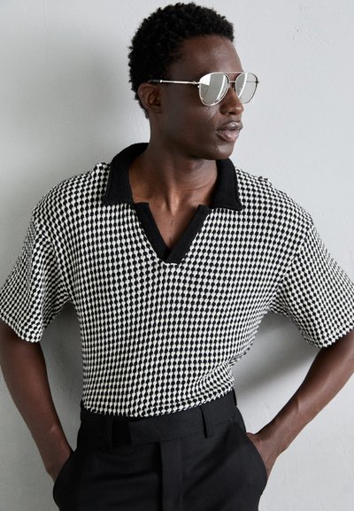 Polo de manga corta a cuadros en blanco y negro con cuello negro de contraste; modelo usando gafas de sol reflectantes y pantalones negros.