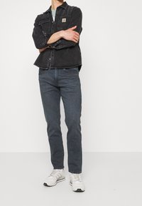 Svart denimjacka med knäppning och två framfickor, kombinerad med mörka jeans och vita sneakers, mot en enfärgad bakgrund.