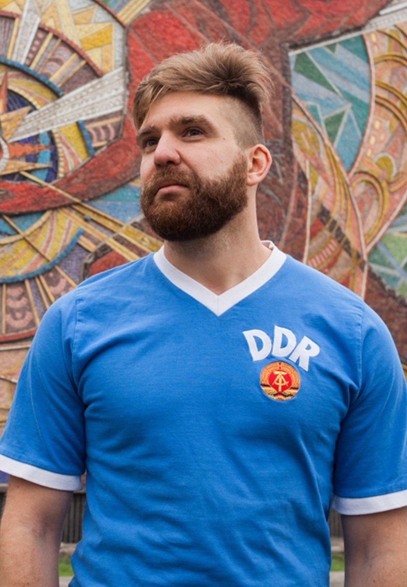 Blaues T-Shirt mit kurzen Ärmeln und weißem V-Ausschnitt, mit "DDR" in Weiß und einem runden Emblem auf rotem Hintergrund. Der Hintergrund hat bunte Muster.