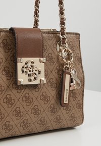 Sac à main en cuir marron motif, avec un rabat en cuir lisse, des accessoires en or, une bandoulière en chaîne et un porte-clés décoratif portant le nom de la marque "Guess."