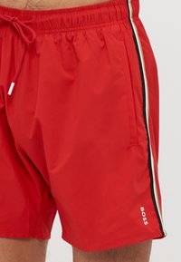 Rote Badekurz-Hosen aus leichtem Stoff, mit einem elastischen Bund, Kordelzug, seitlichen Taschen und kontrastierenden vertikalen Streifen.