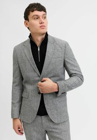 Blazer grigio a fantasia con una vestibilità sartoriale, caratterizzato da una chiusura con un solo bottone, rever a laccio e tessuto testurizzato. Indossato sopra un collare zip nero.