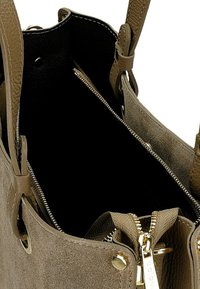 Světlě hnědá semišová taška přes rameno s koženými doplňky. Černý interiér, kapsa na zip a detaily v zlaté barvě. Otevřený vršek s uzlovými popruhy.