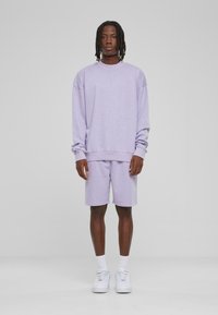 Sweatshirt violet clair avec épaules tombantes et shorts assortis, fabriqué en tissu doux. Des baskets blanches et des poignets côtelés complètent la tenue.