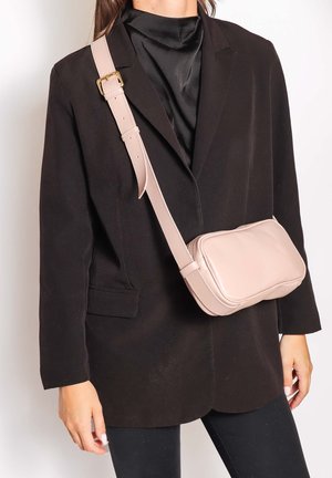 Personne portant un blazer noir et un haut en satin avec un sac bandoulière en cuir beige doté d'une sangle à boucle dorée portée en travers de la poitrine.