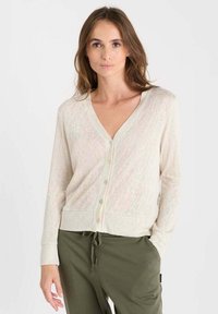 Hellbeige Strickjacke mit V-Ausschnitt, langen Ärmeln und Knöpfen. Hergestellt aus einem strukturierten Stoff, verfügt sie über einen taillierten Bund. Kombiniert mit olivgrünen Hosen.