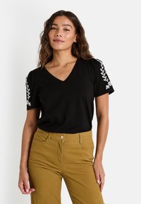 Camiseta negra de cuello en V con mangas abullonadas, que presenta bordados florales blancos en los hombros. Confeccionada con una tela suave y ligera.
