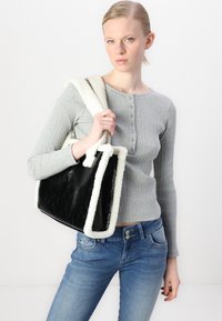 Sac en simili cuir noir avec une bordure en fausse fourrure blanche, de forme rectangulaire, tenu par un mannequin portant un pull à manches longues gris à côtes et un jean bleu.