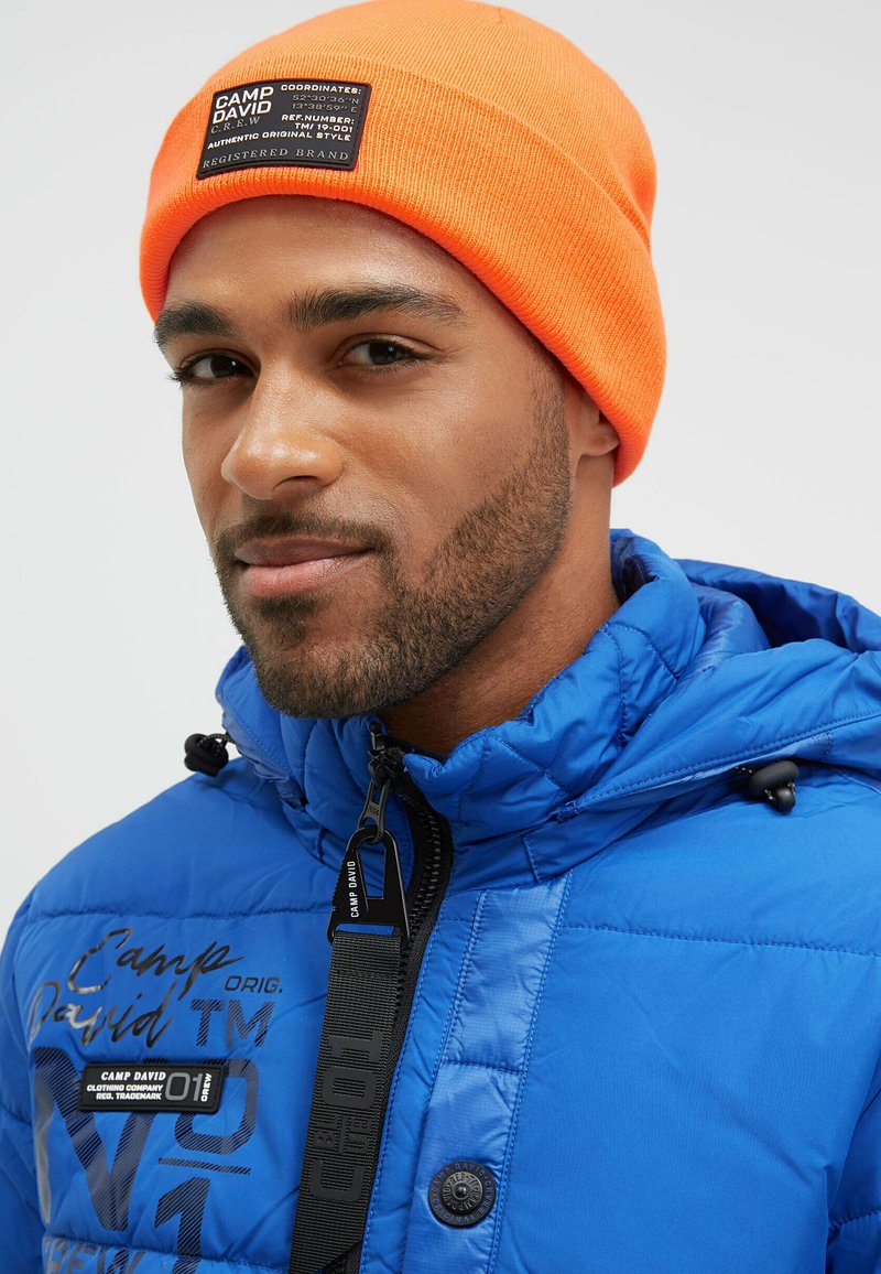 Blue puffer jacket with quilted texture, black zipper accents, and logo patches. Accompanied by a bright orange knit beanie.