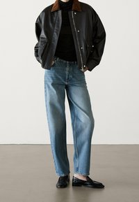 Persoon gekleed in een zwart leren jack met een bruine kraag, een zwarte coltrui, losse blauwe spijkerbroek en zwarte instappers, staand met zijn handen in de zakken.