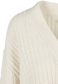 Pull en tricot couleur crème avec col en V côtelé et motif tissé texturé sur l'épaule et le haut de la poitrine.