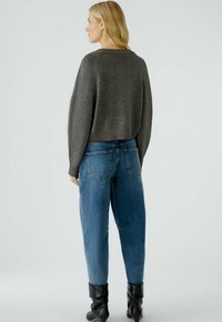 Pull en tricot gris, court, avec un ourlet côtelé et des manches longues, associé à un jean en denim bleu taille haute avec des poches arrière. Des bottes noires complètent le look.