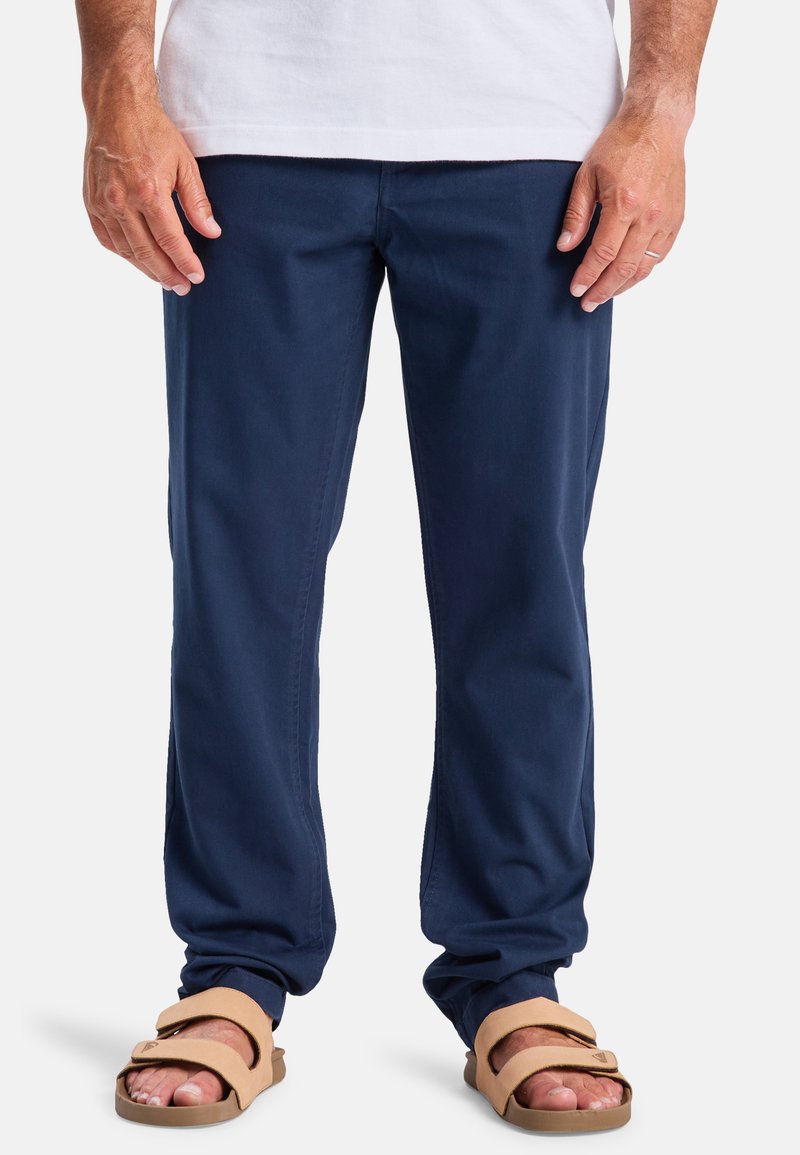 Chinos de algodón azul marino con corte recto, que cuentan con bolsillos frontales y un ligero brillo, combinados con sandalias deslizantes en tono beige.