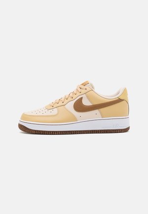 Beige en crèmekleurige Nike Air Force 1 lage sneaker met bruine swoosh en witte tussenzool op een effen witte achtergrond.