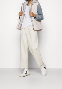 Quiltet vest i beige og denim med hætte i grå, kombineret med cremede bukser og hvide sneakers med sorte detaljer.