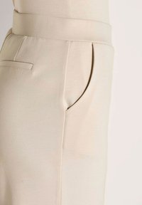 Beige broek met een strakke hoge taille, zijsteekzak en een kleine paspelzak op de achterkant, weergegeven op een neutrale achtergrond.