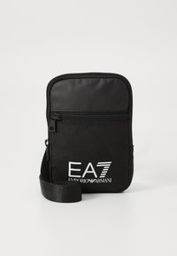 Melnā neilona mazā soma ar rāvējslēdzēju, priekšpusē ar drukātu balto logo "EA7 Emporio Armani" un regulējamu siksniņu.