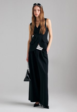 Donna con lunghi capelli castani che indossa un blazer nero senza maniche, pantaloni neri larghi, tacchi neri, occhiali da sole bianchi in testa, che tiene una borsa nera con tracolla a catena.