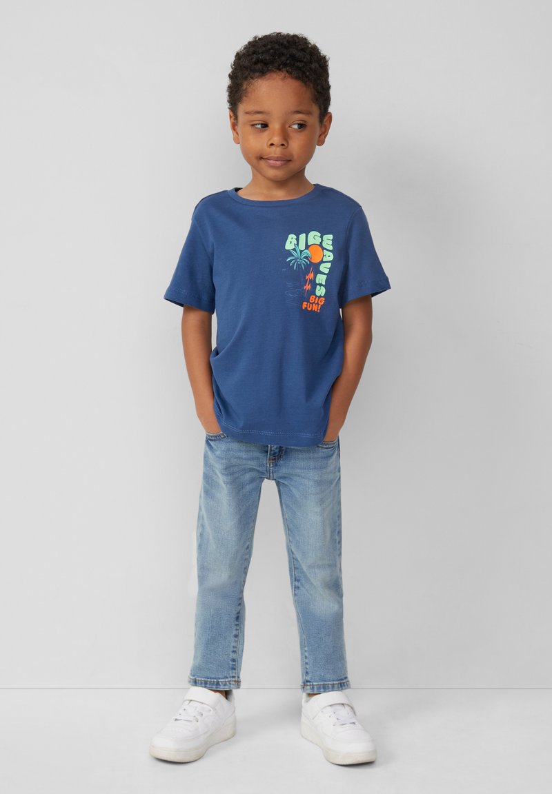 Blaues Baumwoll-T-Shirt mit kurzen Ärmeln, das ein grafisches Design von Wellen, Sonne und Palmen zeigt. Kombiniert mit hellblauen Jeans und weißen Turnschuhen.