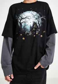 Schwarzes T-Shirt mit einem großen Mond- und Spukhause-Graphic in Hellblau und Gelb. Lange graue Ärmel verlaufen unter dem Shirt.