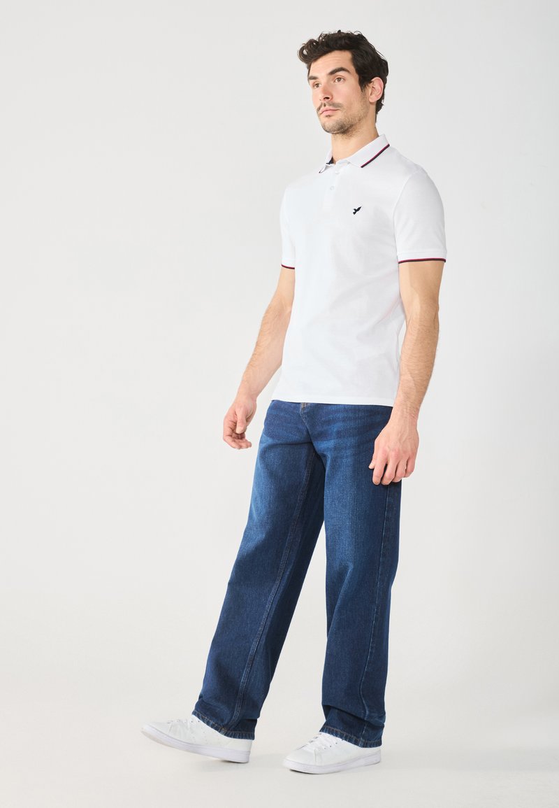 Weißes Poloshirt mit roten Akzenten am Kragen und an den Ärmeln, getragen mit blauen weiten Jeans und weißen Sneakers. Gepflegter, lässiger Look.