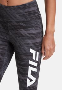 Leggings neri con un design a motivo; presentano un prominente logo Fila bianco sul lato. Il materiale sembra elastico e aderente.