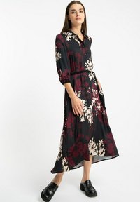 Kobieta stoi z jedną ręką w kieszeni, ubrana w czarną midi sukienkę w czerwony i biały kwiatowy wzór oraz czarne eleganckie loafersy na obcasie.