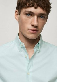 Vaaleanvihreä kauluspaida, jossa on sileä kangas, yksi tasku ja keltaiset napit. Lähikuva paidan designistä.