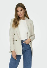 Blazer doppiopetto di un beige chiaro con accenti in bottone dorato, revers a punta e spalle strutturate, abbinato a un top bianco e jeans blu.