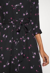 Zwarte jurk met paarse en roze bloemmotieven, knoopsluiting aan de voorkant, strikdetail bij de taille en opgerolde mouwen, gemaakt van lichtgewicht stof.