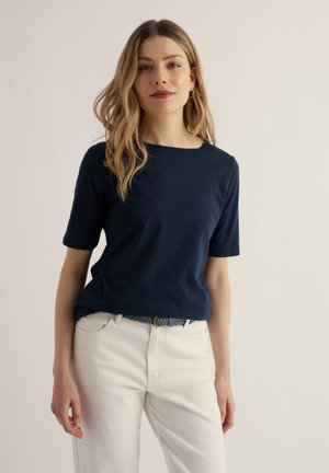 Donna con capelli biondi ondulati che indossa una maglietta blu navy a maniche corte, pantaloni bianchi e una cintura blu intrecciata, in piedi davanti a uno sfondo chiaro.