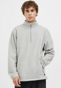 Hellgrauer Fleece-Pullover mit halber Reißverschluss-Kragen, langen Ärmeln und seitlicher Reißverschlusstasche. Mit einem dezenten Logo auf der Brust. Weicher Textil.