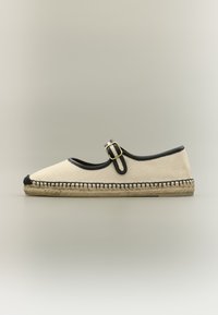 Espadrille plate couleur crème avec bordure noire, une seule bride avec boucle dorée, et semelle en jute tressé sur un fond neutre.