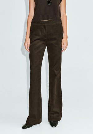 Pantaloni a palazzo in corduroy marrone con righe verticali, vita medio-alta e una consistenza liscia. Abracchiati a un maglione nero a maglia e scarpe nere a punta.