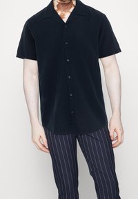 Krátký rukáv, tmavě modrá košile na knoflíky s polosrandou, vyrobená z hladké látky, s uvolněným střihem a jemným leskem. V kombinaci se pruhovanými kalhotami.