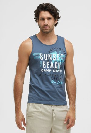 Man with curly dark hair wearing blue tank top with beach text and beige shorts, standing against light gray background.