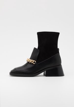Botte noire mi-mollet en cuir et tissu avec talon bloc et détail chaîne dorée sur le devant.